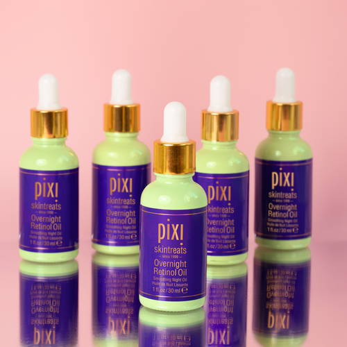 A row of bottles of PIXI Overnight Retinol Oil with purple labels and gold caps, displayed against a pink background.