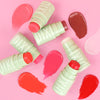 Four Pixi +HydraLip Treat sticks in pale Pixi green packaging arranged on a pink background with corresponding swatches in pink, coral, brown and red.