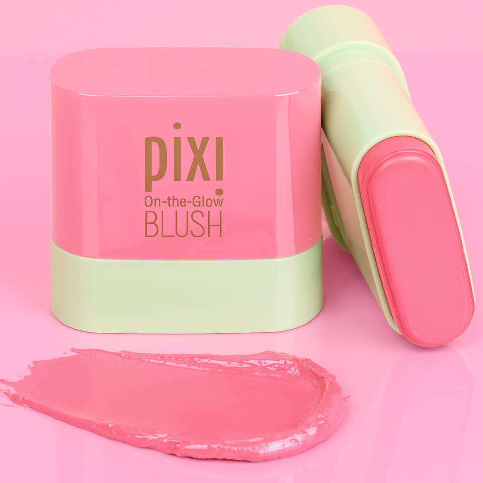 Pixi On-the-Glow Blush Petite stick shown with its pink and mint green cap on, alongside a smear of the pink cream blush on a solid pink background. view 1