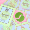 Several Pixi Skintreats DetoxiEYE caffeine and cucumber eye patch packets are arranged on a pink background, with one packet opened to show a green crescent-shaped eye patch in the center.