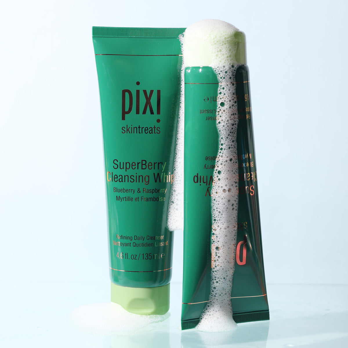 Two Pixi Skintreats SuperBerry Cleansing Whip skincare tubes on a light blue background, one upright showing the SuperBerry Cleansing White product and the other leaning with white lathering foam covering part of the tube.