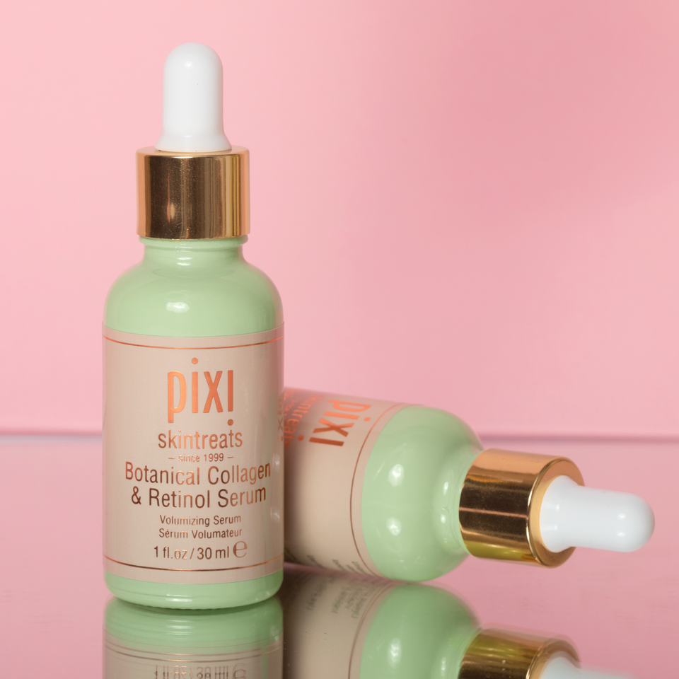 Pixi Skintreats Botanical Collagen & Retinol Serum in a pale mint green glass dropper bottle with a gold collar and white dropper, shown upright with a second bottle lying on its side on a pink reflective surface. view 1