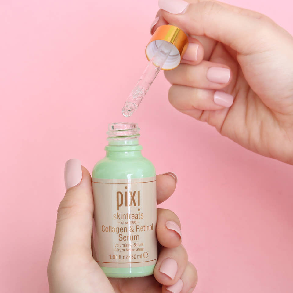 A close-up of a hand holding a Pixi Skin Treats Collagen & Retinol Serum bottle while another hand holds the open dropper above it, with clear serum dripping into the bottle against a solid pink background. view 2