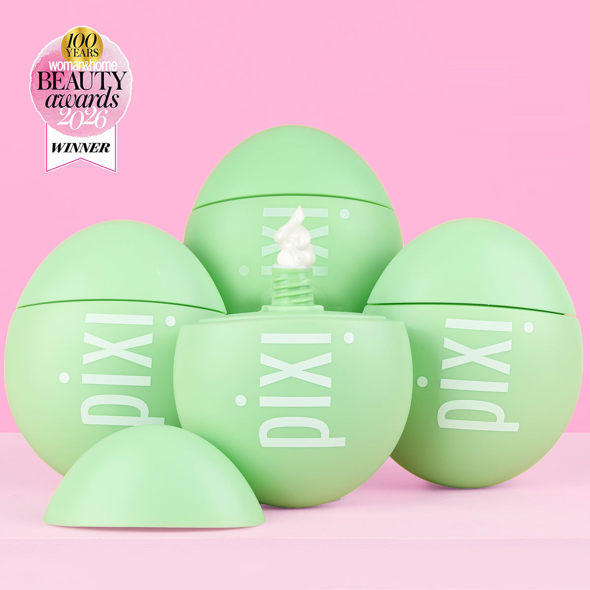 Five light green Pixi cosmetic egg-shaped containers arranged on a pink background, with one open container in the center showing white cream and a small award badge in the top-left corner.
